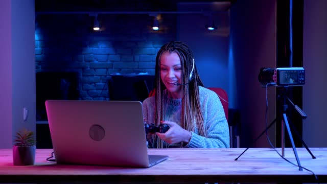 Closeup-shoot-of-young-attractive-female-blogger-with-dreadlocks-in-headphones-playing-video-games-on-the-laptop-with-the-neon-background-indoors