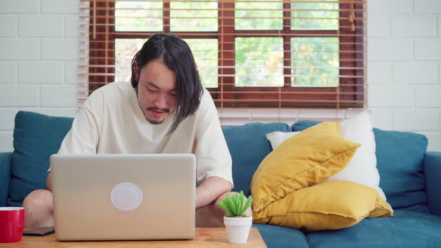 Freiberuflicher-asiatischer-Mann-arbeitet-zu-Hause,-männlich-kreativ-auf-Laptop-auf-dem-Sofa-im-Wohnzimmer.-Business-junge-Mann-Besitzer-Unternehmer,-Computer-spielen,-überprüfen-Social-Media-am-Arbeitsplatz-bei-modernen-Hauskonzept.