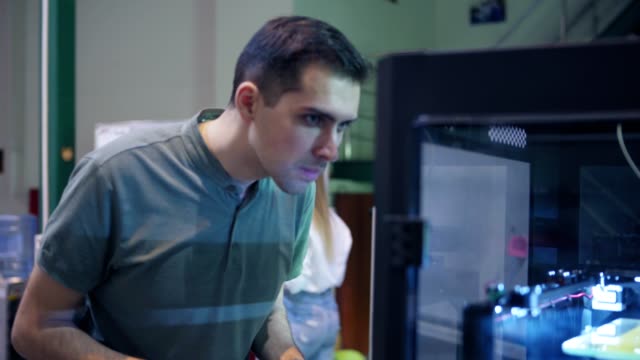 Junger-Ingenieur,-der-im-Labor-auf-eine-3D-Druckmaschine-zugeht,-dreidimensionale-Druckprozesse-beobachtet-und-Notizen-in-seinem-Notizblock-überprüft,-Schuss-verfolgt