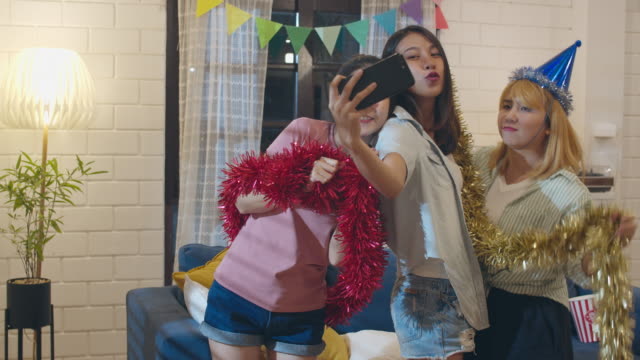 Group-of-Asian-women-party-at-home,-female-using-mobile-phone-making-vlog-selfie-to-social-media-while-funny-dance-together-in-living-room-in-night.-Young-friend-celebrate-holiday-concept.-Slow-motion