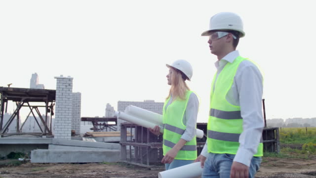 Man-and-woman-supervisor-and-assistant-with-drawings-in-white-helmets-go-and-talk-on-the-construction-site-showing-the-location-of-objects