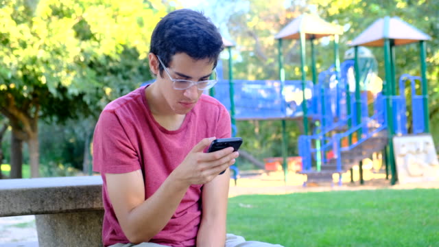 Young-man-using-his-smart-phone-sitting-in-a-park-bench
