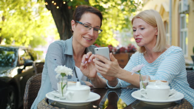 Happy-ladies-laughing-chatting-looking-at-smart-phone-screen-in-outdoor-cafe