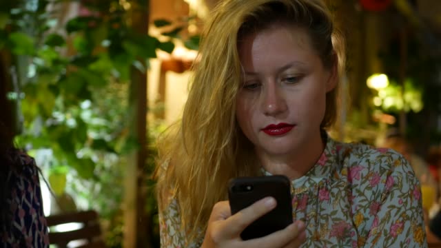 Pensive-girl-uses-a-smartphone,-writes-a-message,-chatting.-Woman-looking-for-information-in-the-phone-and-sad.-Sitting-in-restaurant-alone.-Evening-time,-lowlight.-Technology-and-entertainment-concept.-4k