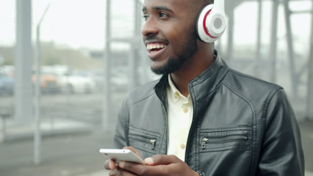 Afroamerikanischer-Hipster-in-Kopfhörern-berührt-Smartphone-Bildschirm-draußen
