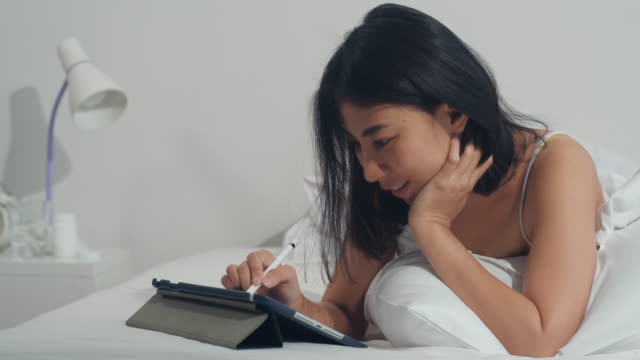 Young-Asian-woman-using-tablet-checking-social-media-feeling-happy-smiling-while-lying-on-bed-after-wake-up-at-house-in-the-morning,-Attractive-indian-female-smiling-relax-in-bedroom-at-home-concept.