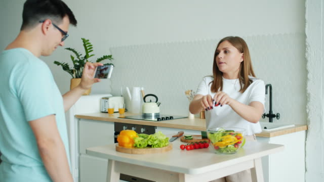 Mädchen-Kochen-Salat-sprechen-zeigt-Daumen-up,-während-Kerl-Aufnahme-Video-für-vlog