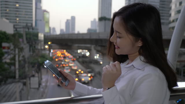 Junge-lächelnde-Geschäftsasiatische-Frau-mit-Smartphone-in-der-modernen-Innenstadt-in-der-Dämmerung-von-Bangkok-Thailand.-Sie-benutzt-ihren-Finger,-um-den-Bildschirm-zu-berühren.-Zum-Tragen-von-Hologrammen-entwerfen-Sie