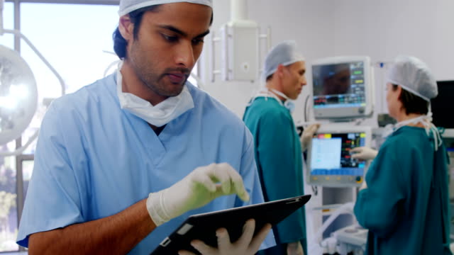 Cirujano-usando-tableta-digital-mientras-sus-colegas-interactúan-en-segundo-plano