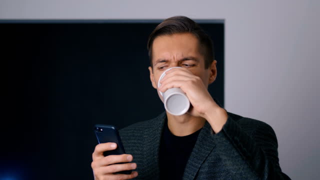 Fokussierter-und-stirnrunder-Mann-Unternehmer-trinkt-Kaffee-und-verwendet-ein-Smartphone-im-Büro-während-einer-Kaffeepause.