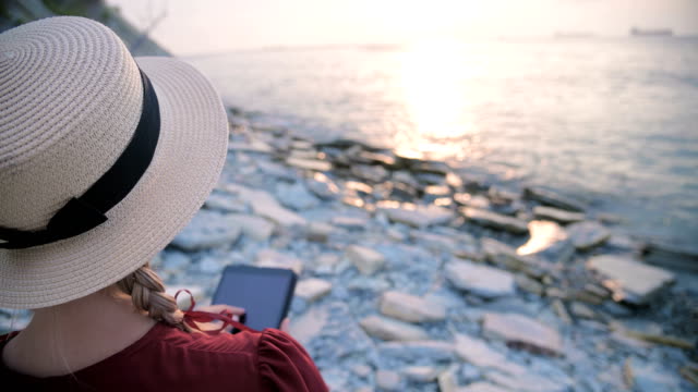 Close-up-View-from-the-back-An-attractive-young-girl-in-a-summer-red-dress-and-a-straw-hat-sits-on-a-stone-by-the-sea-at-sunset-and-watches-something-on-a-tablet.-Swipe-across-the-screen