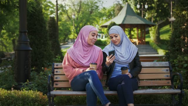 Dos-mujeres-musulmanas-de-piel-oscura-en-coloridos-hiyabs.-Se-ríen,-hablan,-miran-la-pantalla-del-teléfono-inteligente.-Sentado-en-el-banco-del-parque.-"Lose-up"