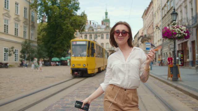 The-girl-is-going-and-listening-to-music-with-headphones.-She-is-very-happy-and-dancing.-Tram-is-riding-and-people-are-walking-in-the-background.-4K