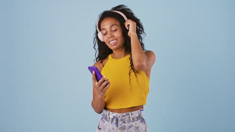 Afro-american-female-is-smiling,-dancing,-listening-to-the-music-through-headphones-and-looking-at-her-cellphone.-Posing-on-blue-background