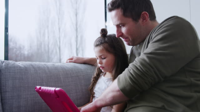 Vater-und-Tochter-sitzen-auf-Sofa-zu-Hause-spielen-zusammen-auf-digitalen-Tablet-in-rosa-Fall-zu-Hause
