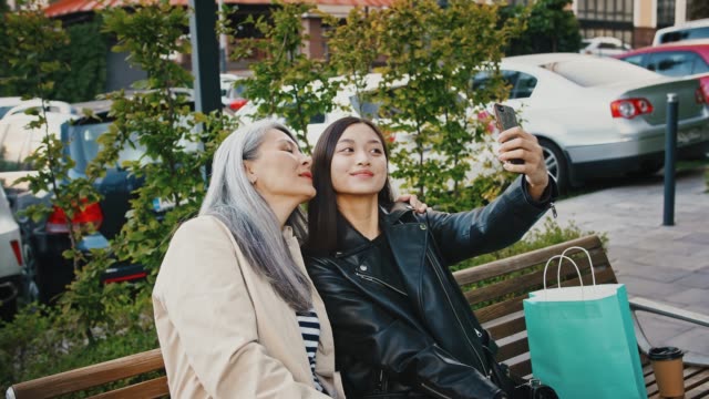 Asian-mature-woman-and-her-daughter-are-smiling-and-showing-victory-sign,-taking-selfie-by-smartphone-while-sitting-outdoors-on-a-bench