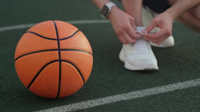 Unbekannter-Sportler-bindet-Schnürsenkel-an-Turnschuhen,-die-in-der-Nähe-eines-Basketballballs-posieren