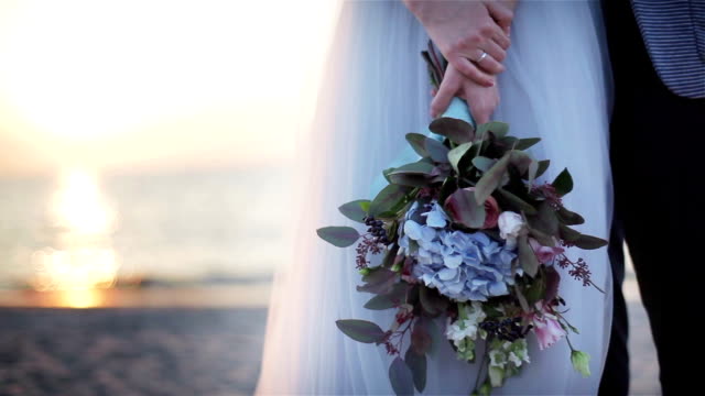 Unusual-wedding-bouquet-close-up-outdoors.-Woman-hands-hold-bridal-bouquet-made-from-flowers-and-green-leaves-at-sunset-sea-view-background.-Floral-design-decoration.-Newlyweds-posing-at-beach-no-face