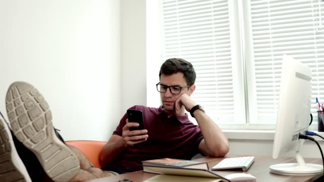Young-worker-in-glasses-relaxing-with-his-legs-on-the-table-modern-office-using-phone.-Bored-manager-surfing-in-internet.-Shot-4k