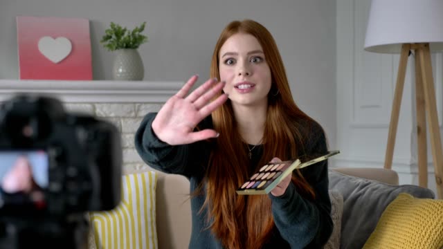 Young-red-haired-girl-blogger,-smiling,-talking-at-the-camera,-showing-a-new-purchase,-cosmetics,-Eyeshadow-Pallet,-home-comfort-in-the-background.-60-fps