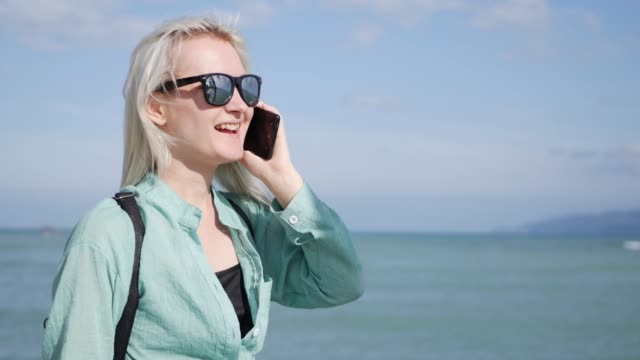 Beautiful-slim-woman-with-long-blonde-hair-and-green-shirt-standing-and-talking-on-phone-over-background-sea
