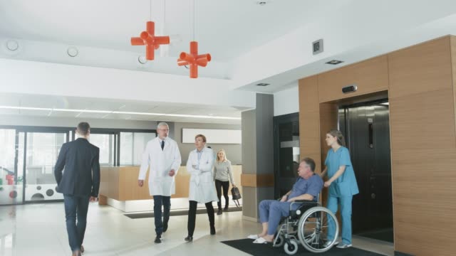 First-Floor-of-the-Busy-Hospital,-Doctors,-Nurses-and-Personnel-Busy-Working,-Receptionist-Talks-with-a-Patient,-Assistant-Moves-Elderly-Man-in-the-Wheelchair.-New-Modern-Medical-Hospital-with-Professional-Staff.