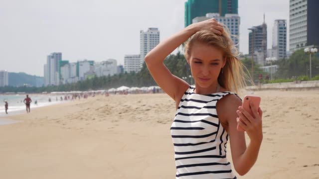 Young-beautiful-slim-woman-with-long-blonde-hair-in-black-and-white-dress-making-selfie-on-mobile-phone-near-the-sea-on-background-of-city