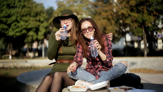 Dos-mujeres-riendo-y-cantando-en-el-parque-durante-el-almuerzo.-Dos-chicas-hipster-comiendo-bocadillos-en-el-parque,-reír-y-hablar.-Estancia-en-el-parque.-El-sándwich-de-vacaciones.