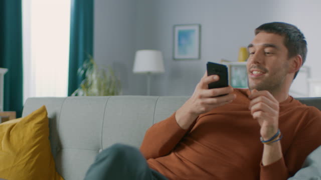 Gut-aussehend-glücklicher-Mann-zu-Hause-sitzt-unten-auf-dem-Sofa-während-der-Verwendung-von-Smartphone,-hat-verschiedene-berühren-und-Swiping-Gesten.-Mann-mit-Handy-in-seinem-gemütlichen-Wohnzimmer-entspannen.