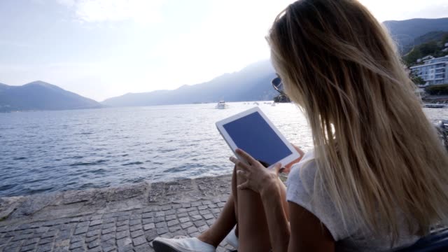 Young-woman-sitting-on-a-bench-in-a-town-facing-the-lake-and-mountains-using-a-digital-tablet-technology--Mobile-apps,-online-reservation-concept