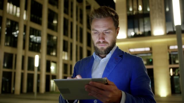 Medium-shot-of-serious-businessman-using-tablet-computer-in-illuminated-might-street