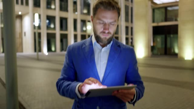 Tilt-down-medium-shot-of-serious-businessman-browsing-Internet-on-tablet-computer-while-walking-in-night-city-street