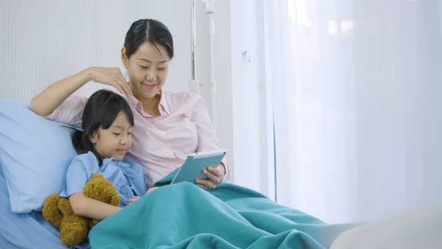 Cute-little-girl-lying-on-bed-with-her-mother-in-hospital,-watching-funny-cartoons,-movies-on-digital-tablet.-illness-and-treatment.