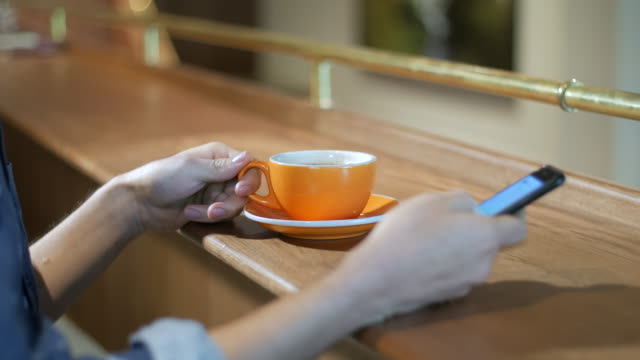 Nahaufnahme-von-den-Händen-eines-Mannes-sitzt-an-einem-Tisch-Handy-halten-und-trinken-Kaffee-aus-einem-orange-Cup,-überprüfen-von-Nachrichten,-chatten-in-sozialen-Medien