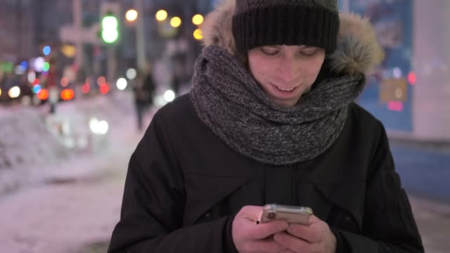 Man-walking-on-street-in-warm-clothing-and-text-messaging-on-mobile-phone-at-night