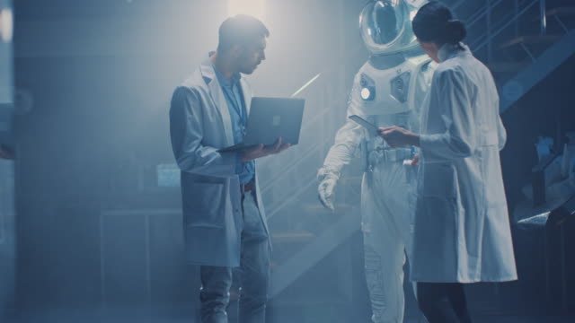 Diverse-Teams-von-Luft-und-Raumfahrt-Wissenschaftlern-und-Ingenieuren,-die-weiße-Mäntel-tragen,-haben-Diskussionen,-Benutzung-von-Computern,-Design-New-Space-Suit,-die-für-Galaxy-Exploration-und-Reisen-angepasst-sind.-Astronaut-bauen-Anzug