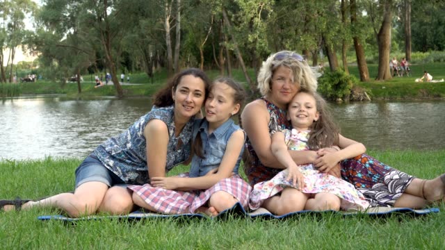 Same-sex-family-with-children-playing-and-resting-in-nature.-Two-mothers-with-young-daughters-hugging-and-having-fun,-sitting-on-the-lawn-in-the-Park-at-sunset-on-a-summer-day-on-the-river-Bank.-4K.
