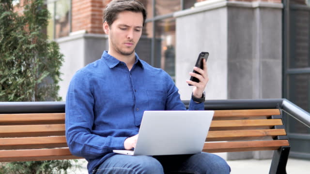 Hombre-joven-usando-smartphone-y-laptop,-sentado-en-el-Banco
