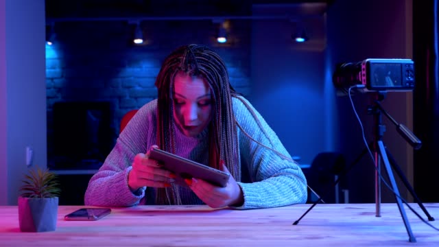 Closeup-shoot-of-young-attractive-female-blogger-with-dreadlocks-playing-video-games-on-the-tablet-getting-emotional-streaming-live-live-with-the-neon-background-indoors