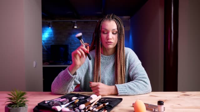 Closeup-shoot-of-young-attractive-female-makeup-artist-with-dreadlocks-streaming-live-and-advertising-cosmetics-with-the-neon-background-indoors