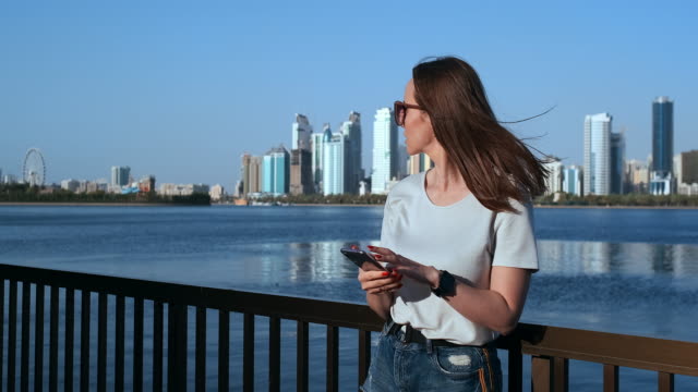 Hermosa-chica-con-el-pelo-largo-en-gafas-de-sol-usando-aplicación-de-teléfono-inteligente-en-Sunset-River-Quay-cerca-de-4K.