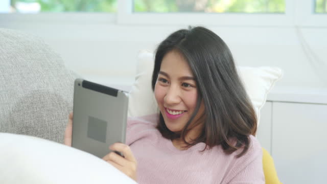 Asian-woman-listening-music-and-using-tablet,-female-using-relax-time-lying-on-home-sofa-in-living-room-at-home.-Happy-female-listening-music-with-headphones-concept.