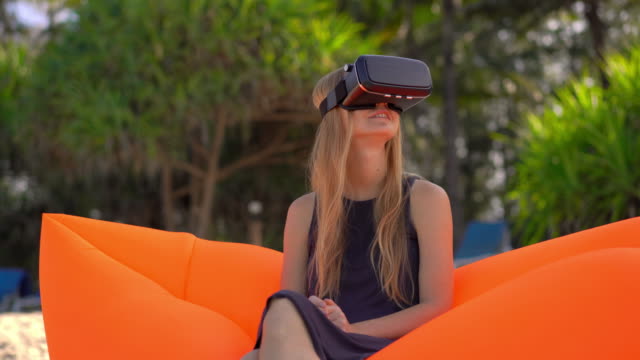 Junge-Frau-sitzt-auf-einem-aufblasbaren-Sofa-an-einem-tropischen-Strand-mit-einer-VR-Brille.-Konzept-moderner-Technologien,-die-Ihnen-das-Gefühl-geben-können,-woanders-zu-sein