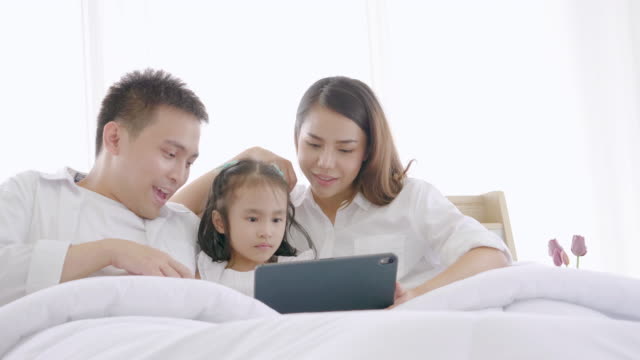 Asiatische-Familie-lacht-und-schaut-in-digitalen-Tablet,-während-auf-dem-Bett-im-Schlafzimmer-liegen