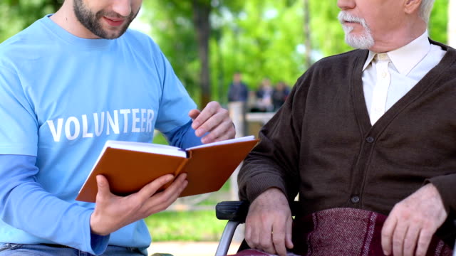 Male-volunteer-reading-book-to-lonely-disabled-pensioner-in-park,-social-support