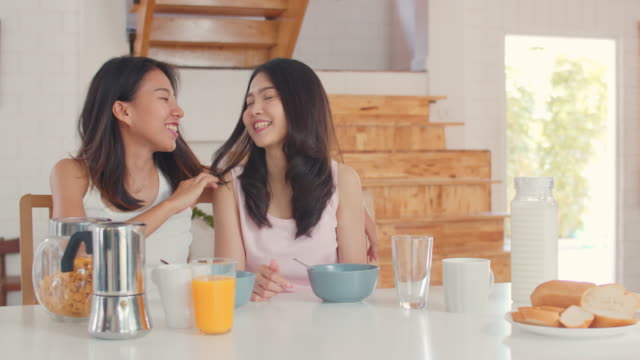 Asian-Lesbian-couple-has-breakfast-drink-juice,-cereal-and-milk-in-bowl-on-table-in-kitchen-at-home.
