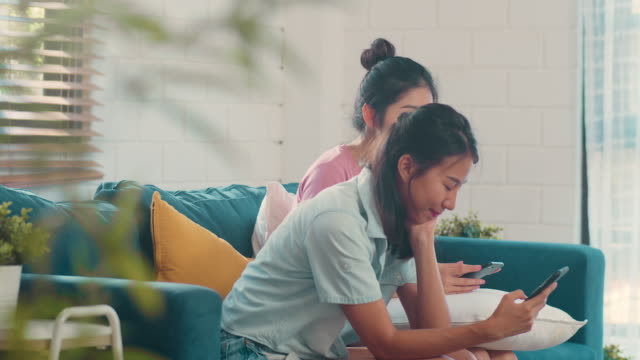 Junge-asiatische-Lesbische-Frauen-Paar-mit-Handy-überprüfen-Social-Media-im-Internet-zusammen,-während-liegen-Sofa-im-Wohnzimmer.