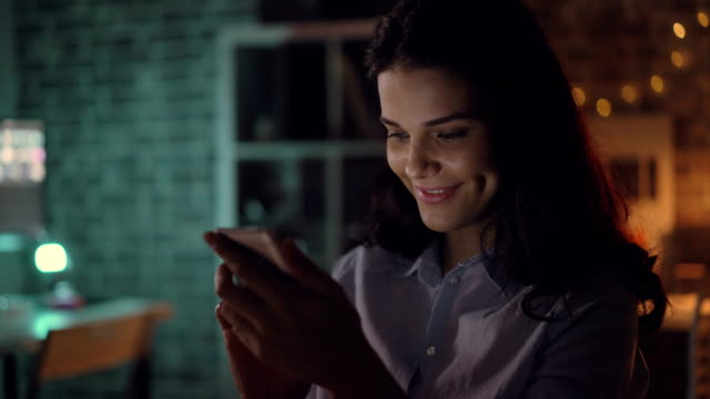 Cheerful-young-woman-using-smartphone-at-night-in-dark-office-smiling