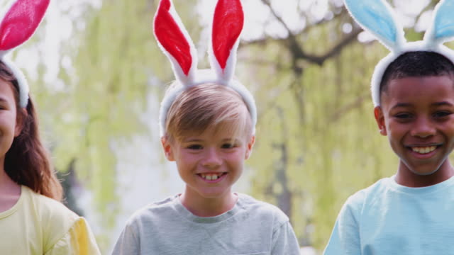 Portrait-of-group-of-children-wearing-bunny-ears-on-Easter-egg-hunt-in-garden-smiling-at-camera---shot-in-slow-motion