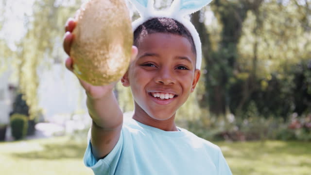 Portrait-of-boy-wearing-bunny-ears-on-Easter-egg-hunt-outdoors-at-home-holding-up-chocolate-egg-to-camera-and-smiling---shot-in-slow-motion